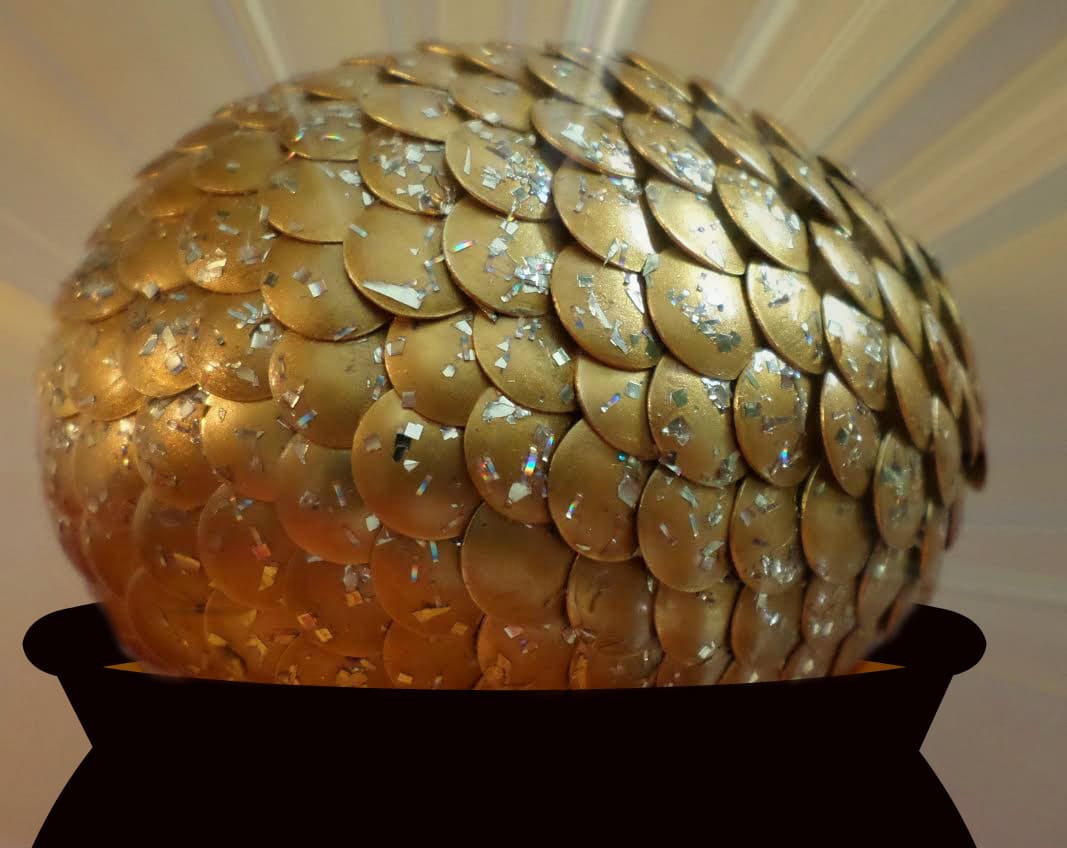 A large golden egg covered with shiny, overlapping sequins sits in a black cauldron, with rays of light radiating from behind it.