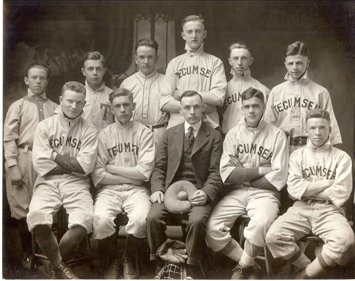 A vintage black-and-white photo of a baseball team with ten young men in Tecumseh uniforms and one man in a suit seated in the center, holding a baseball, posing for a formal team portrait.