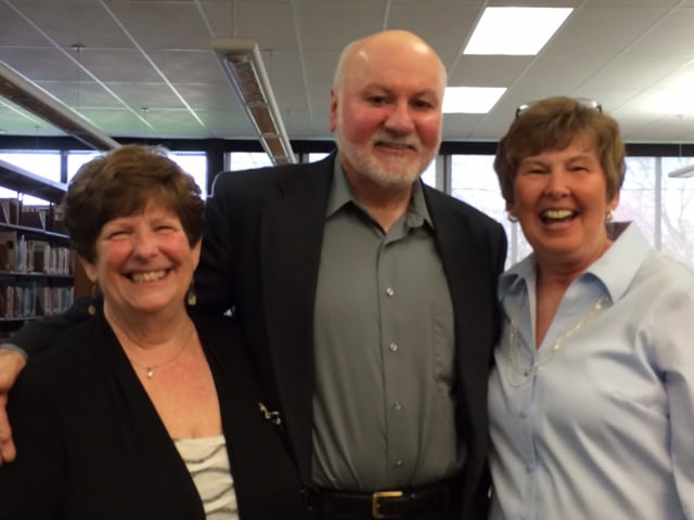 Three older adults, two women and one man, stand close together smiling in a library setting with bookshelves and large windows in the background.