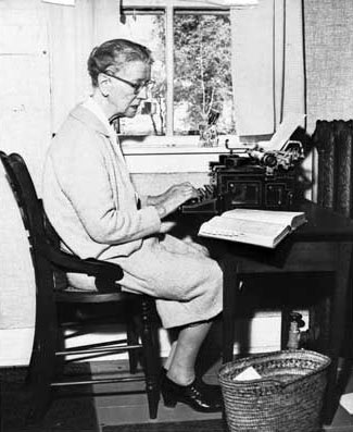 An elderly woman wearing glasses and a light-colored dress sits at a desk, typing on a typewriter beside an open book. She is next to a window, with a basket on the floor by her feet.