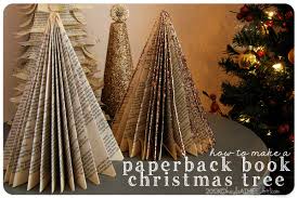 Three Christmas tree decorations, including two made from folded book pages and one glittery cone, are displayed on a table next to a decorated Christmas tree. Text reads: how to make a paperback book Christmas tree.