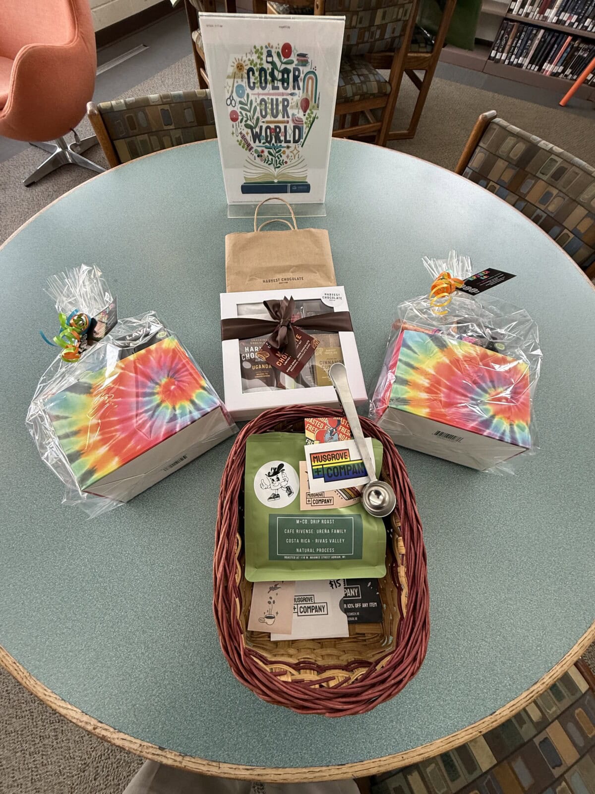 A round table displays colorful gift baskets, tie-dye wrapped boxes, a sign reading Color Your World, and a wicker basket with books, treats, and a mug in a library setting.