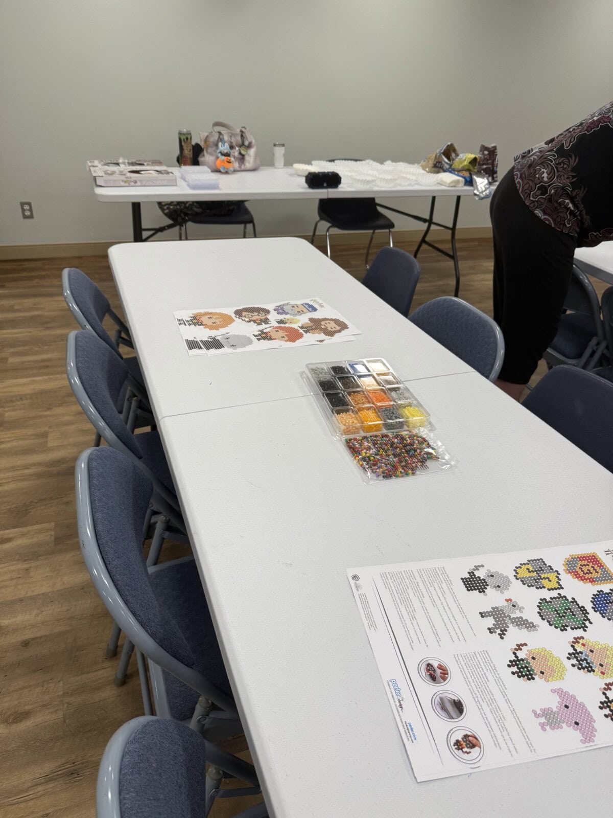 A white table with craft supplies, including beads and pattern sheets, is surrounded by blue chairs. Another table with various items and a person partially visible is in the background in a room with wooden floors.