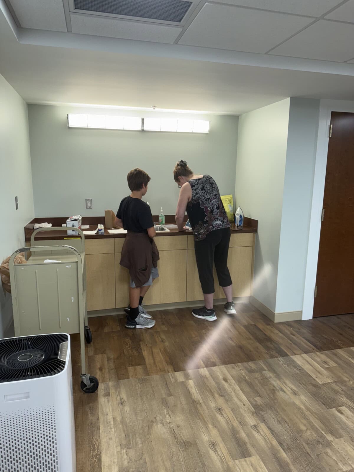 A woman and a boy stand at a counter in a brightly lit room with wood floors. The woman is leaning over the counter, and the boy stands beside her. Nearby are a trash can, a cart, and a closed wooden door.