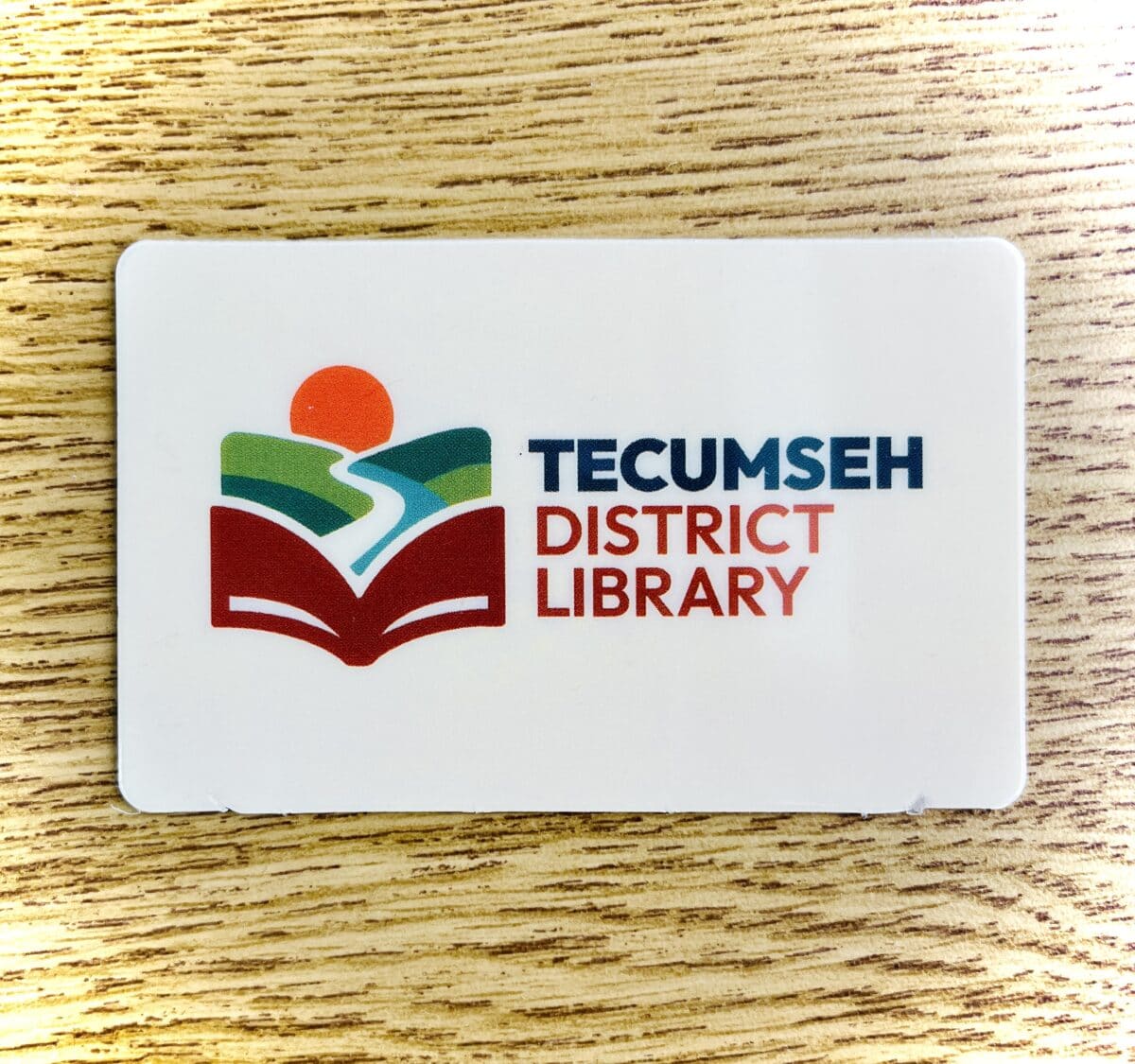 A Tecumseh District Library card on a wood surface, featuring a logo with a red book, green fields, a blue river, and an orange sun above, next to the librarys name in blue and red text.