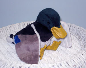 A plush toy duck with yellow beak and feet, green head, and brown, white, and blue body sits on a white wicker surface against a light blue background.