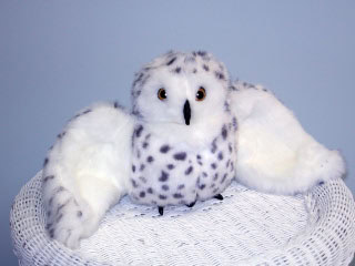 A plush toy snow owl with outstretched wings, white with black spots, sits on a white wicker basket against a pale blue background.