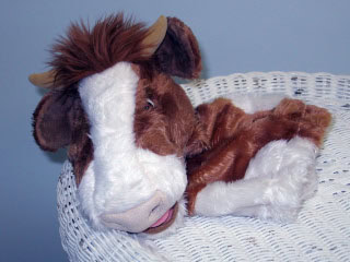 A brown and white cow puppet with soft fur lies on a white wicker surface against a plain light blue background.