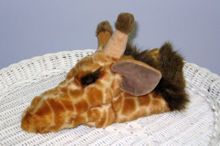 A plush giraffe head with brown spots, tan horns, and large brown ears rests on a white wicker surface against a light gray background.