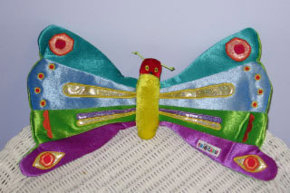 A colorful plush butterfly toy with blue, green, yellow, red, and purple wings, decorated with shiny and patterned fabric, sits on a white woven surface against a light purple background.