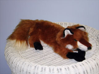 A plush toy fox with reddish-brown fur and a white-tipped tail lies on a round, white wicker basket against a pale blue background.
