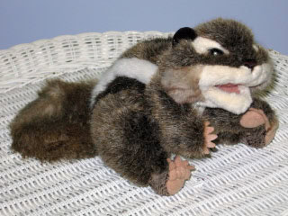 A plush toy resembling a chipmunk or squirrel with brown fur, black and white markings, and pink paws, sits on a white wicker surface.