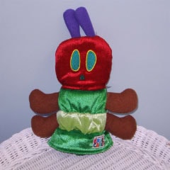 A plush toy resembling the Very Hungry Caterpillar, featuring a red head with green and yellow eyes, brown legs, a green body, and two purple antennae, sitting on a white wicker surface.