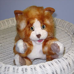 A plush toy cat with orange and white fur sits on a white wicker surface, looking forward with wide eyes and an open mouth.