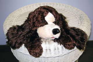 A brown and white stuffed dog toy is lying on top of a round, white wicker basket against a plain background.