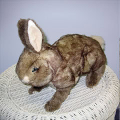 A brown plush rabbit toy with white inner ears and a white tail is standing on a white, woven wicker surface against a light purple background.
