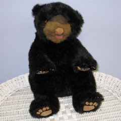 A black plush bear with brown snout and paws is sitting upright on a white wicker surface against a light blue background.