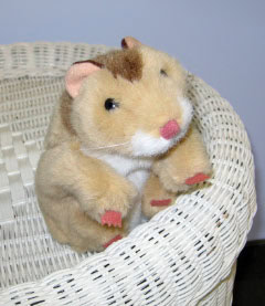 A plush toy hamster with light brown fur, pink nose, and feet sits on the edge of a white wicker basket against a plain background.