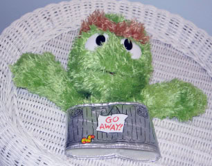 A green, fuzzy puppet with brown hair and big eyes sits in a silver trash can labeled GO AWAY! in red letters, placed on a white wicker chair.