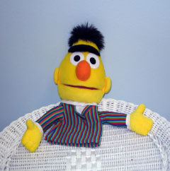 A yellow puppet with a unibrow, orange nose, and striped shirt sits on a white wicker chair against a light blue background.